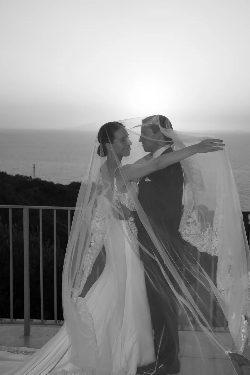 Black and white photo of the couple on the terrace, the veil lifted by the wind.