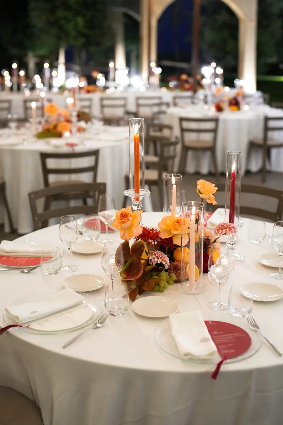 Tavolo rotondo con fiori arancioni, candele e un menù rosso a ventaglio.