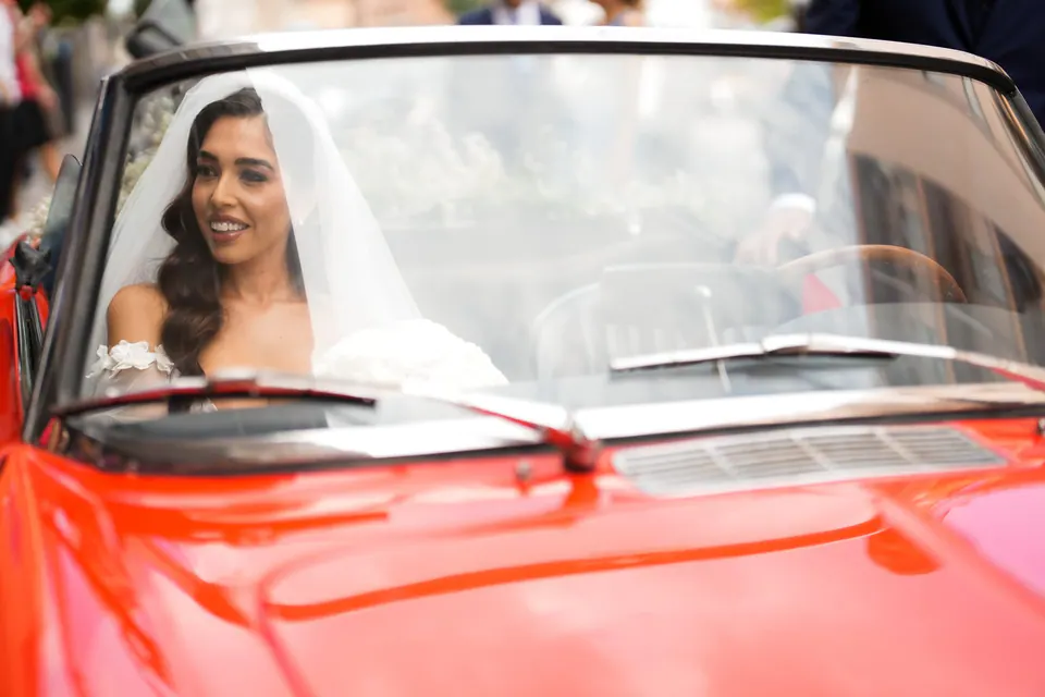 Bride smiling in the red vintage convertible, her veil over her shoulders.