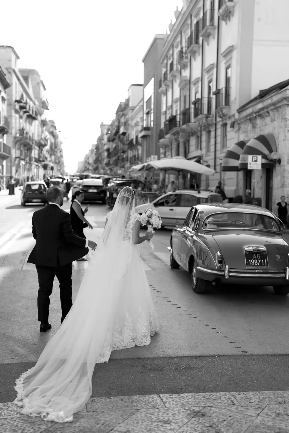 Black and white photo of the bride with her long veil in a street among parked cars
