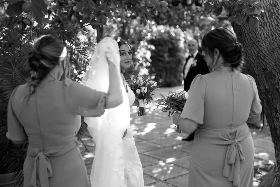 Black and white photo of the bridesmaids fastening the bride's gown outdoors.