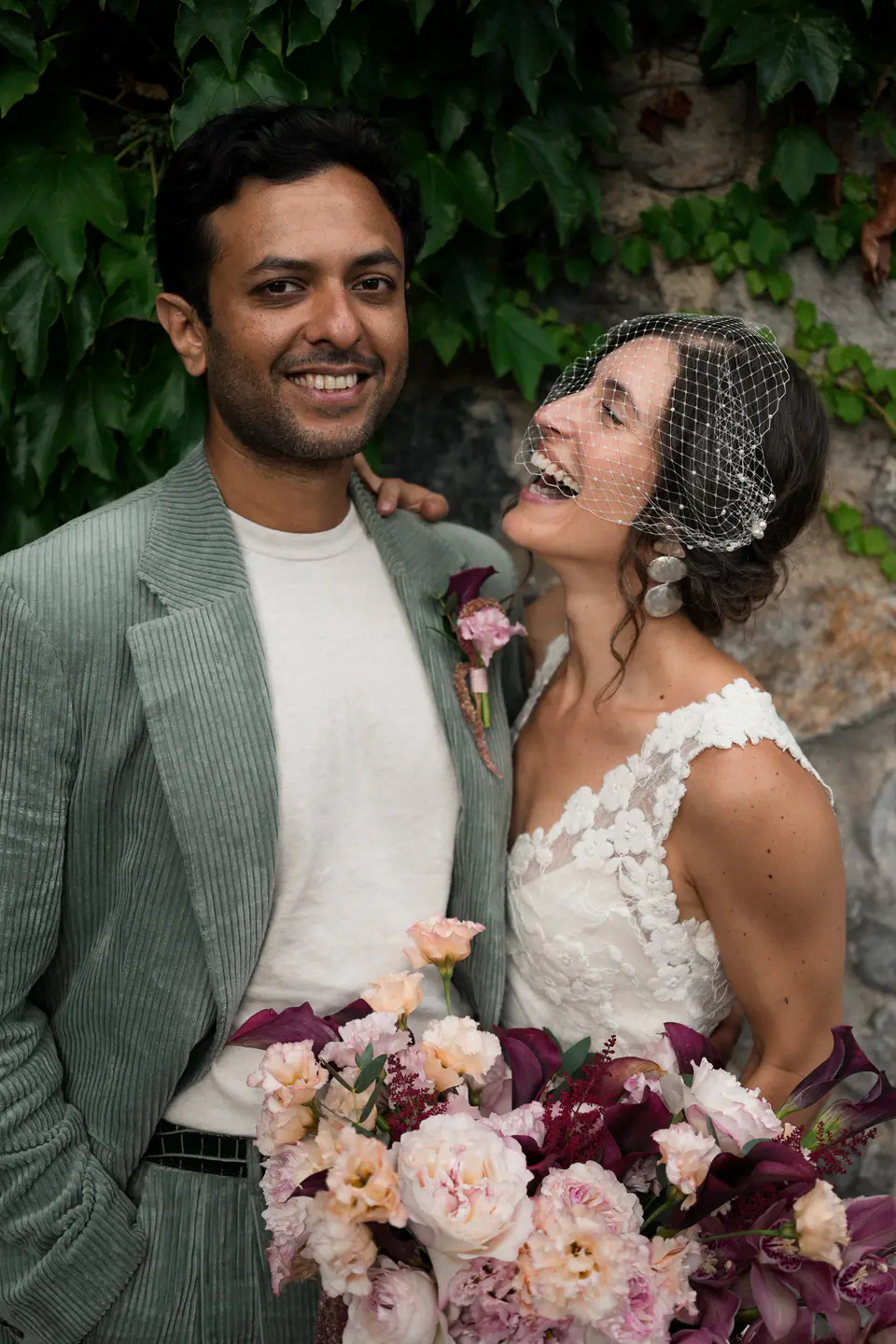 Groom laughing as the bride looks up at him, ivy on the wall behind.