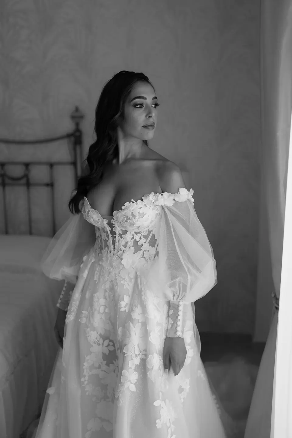 Black and white photo of the bride standing in the bedroom in her off-the-shoulder gown.