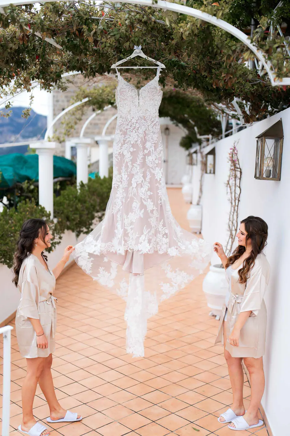 Wedding gown hanging from the pergola on the villa terrace as the bride and a bridesmaid look on.