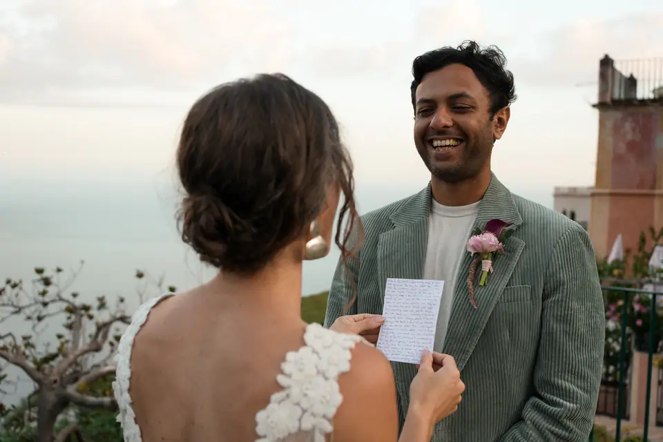 Bride reading her vows from a handwritten letter, the groom smiling.