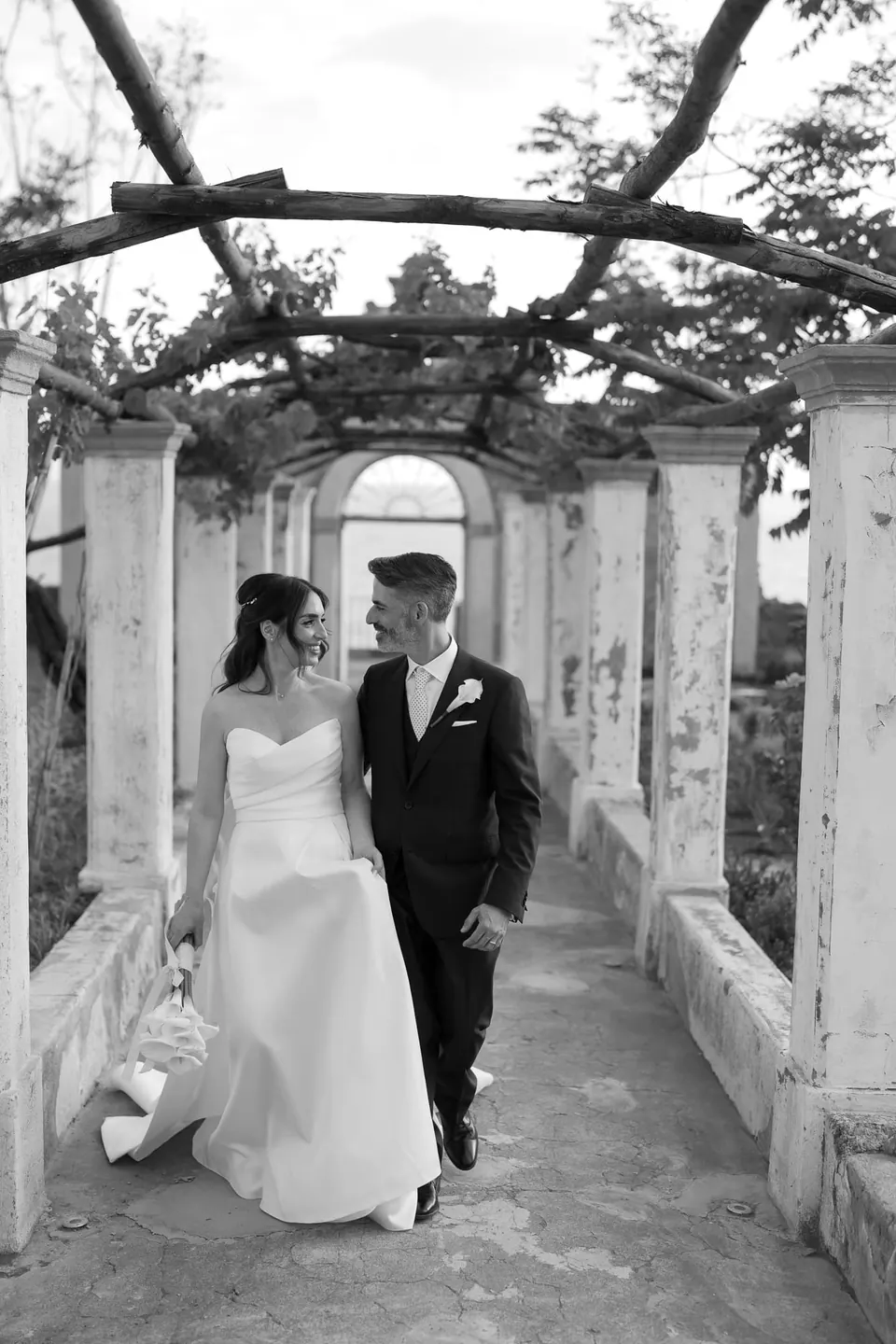 Foto en blanco y negro de los novios caminando bajo una pérgola.
