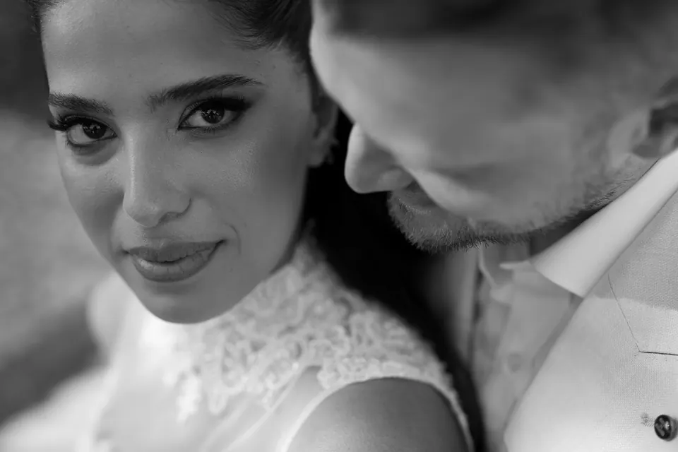 Gros plan en noir et blanc de la mariée qui regarde l’objectif, le visage du marié contre sa joue.
