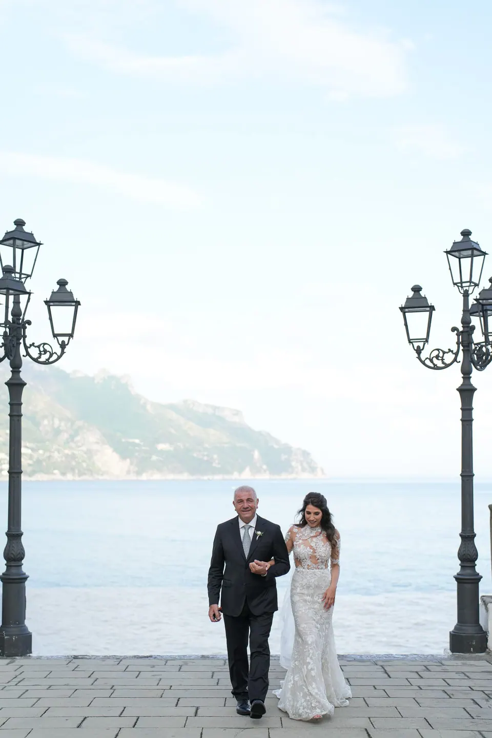 Die Braut geht mit ihrem Vater zwischen Laternen über dem Meer.