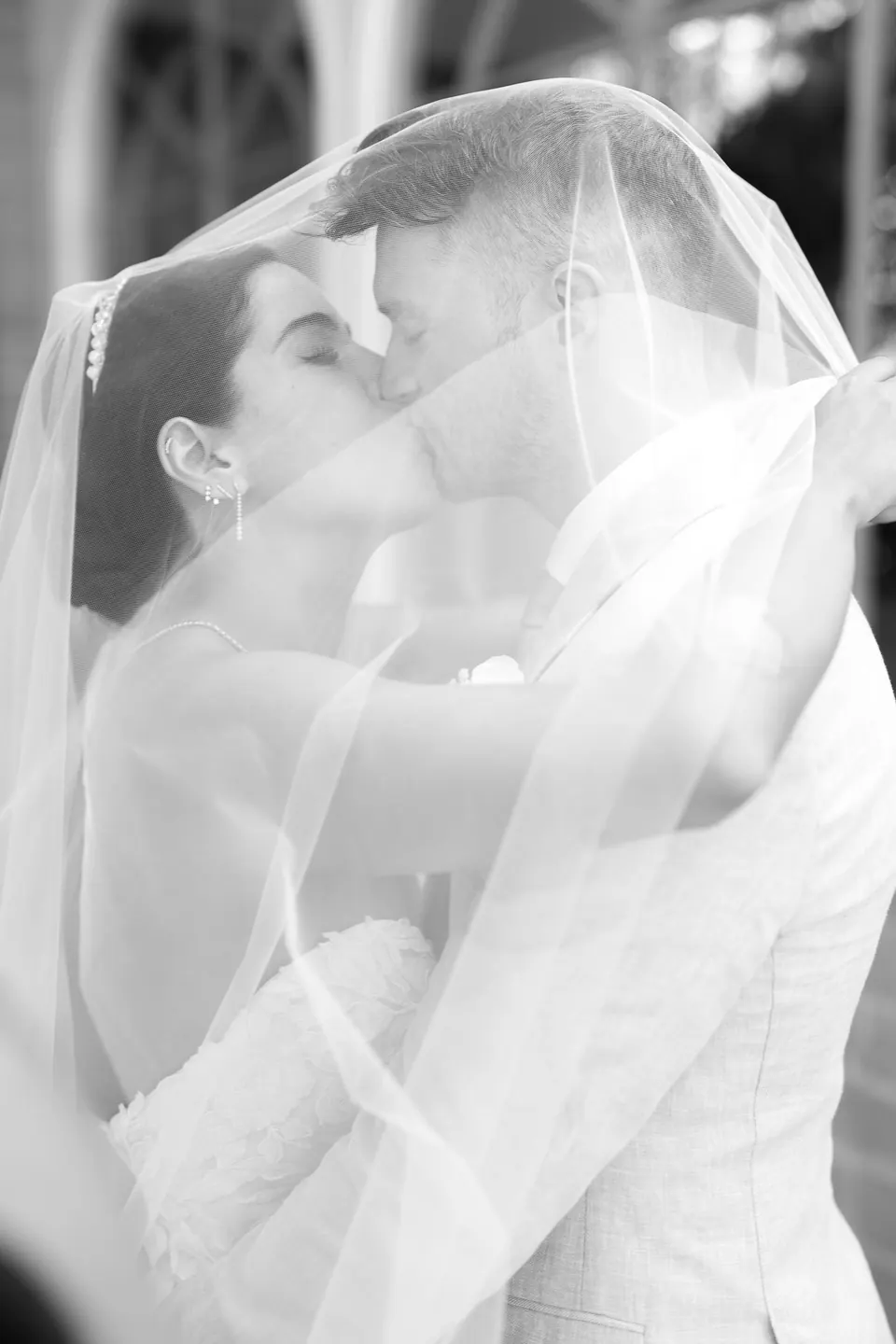 Les mariés s’embrassent sous le voile de la mariée, en noir et blanc.