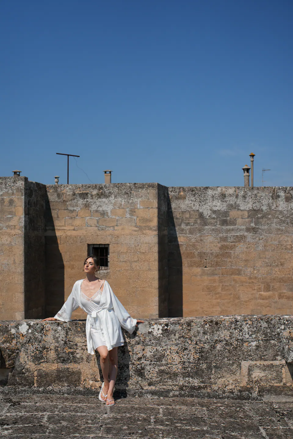 La sposa in vestaglia seduta su un antico muro di pietra sotto un cielo blu intenso.
