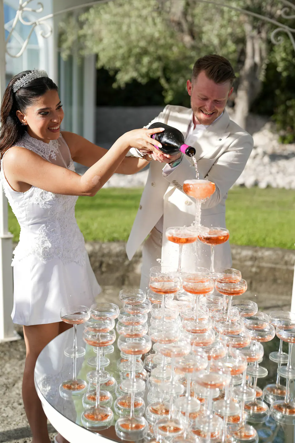 Les mariés versent du rosé dans une pyramide de coupes sur une table miroir du jardin.