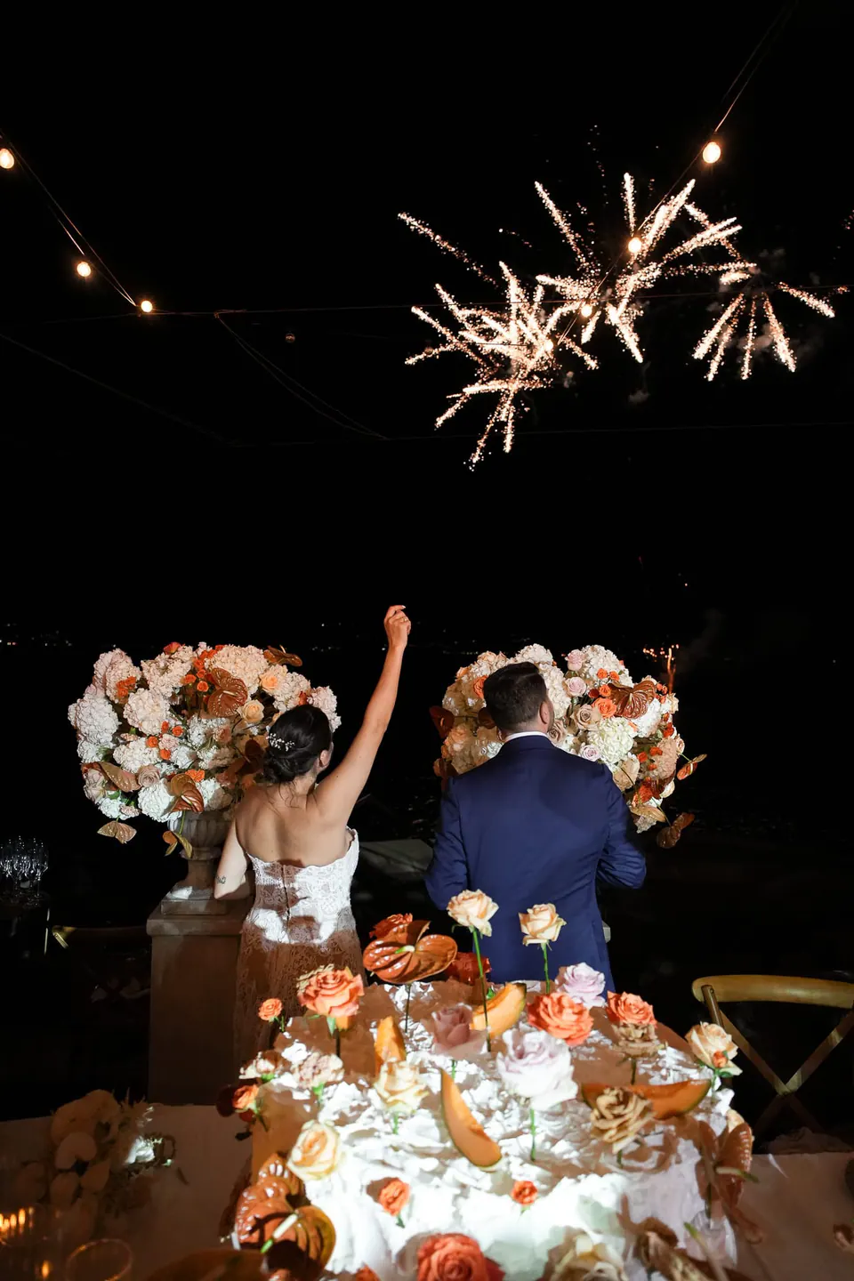 Das Brautpaar erhebt hinter der Torte die Gläser, während darüber Feuerwerk explodiert.