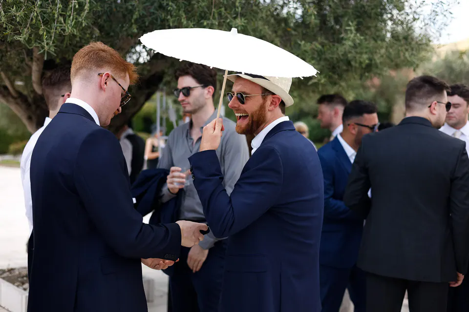 Un invité sous une ombrelle en papier blanc rit avec des amis en costume bleu marine.