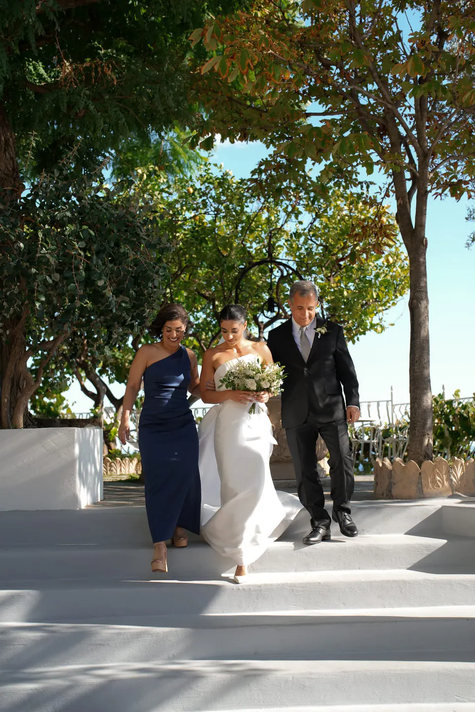 Bride walking to the ceremony with a bridesmaid and her father under the trees.