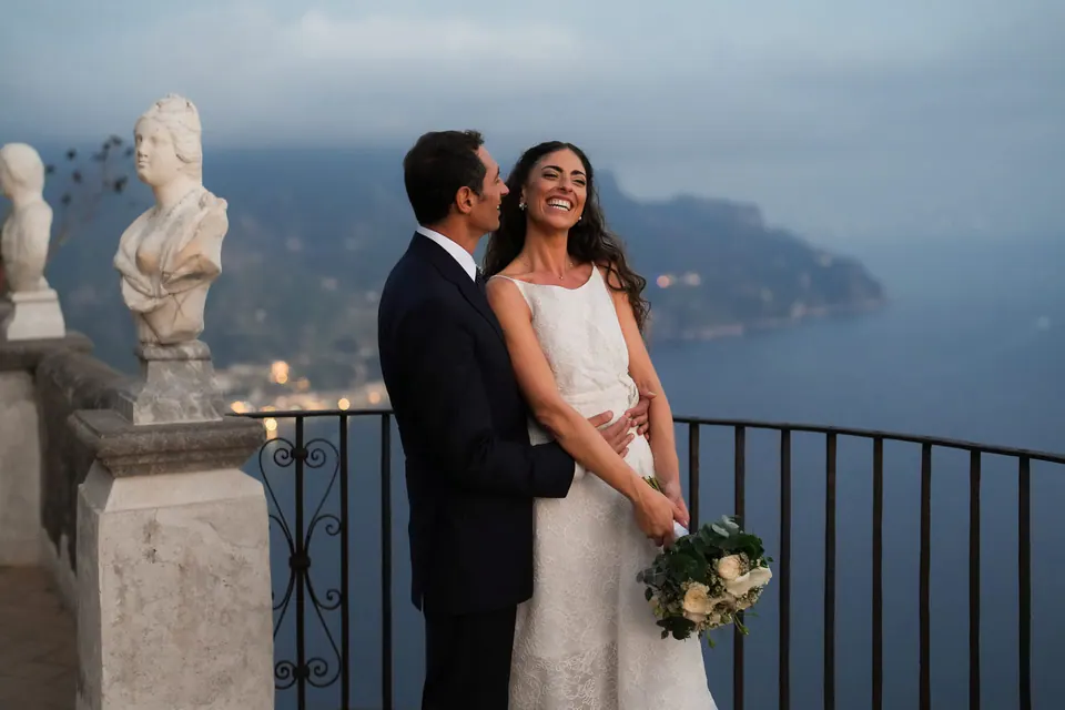 Bride and groom smiling beside a marble bust on the Terrace of Infinity, the coast at dusk.