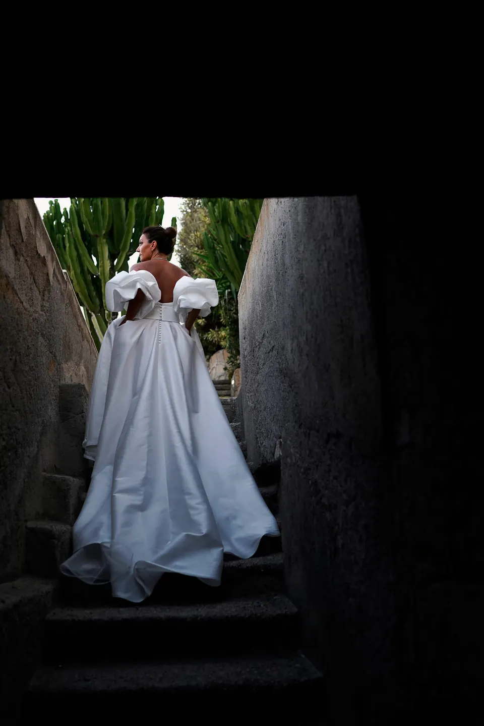 Bride walking down old stone steps in her off-the-shoulder gown, framed by the dark passage.
