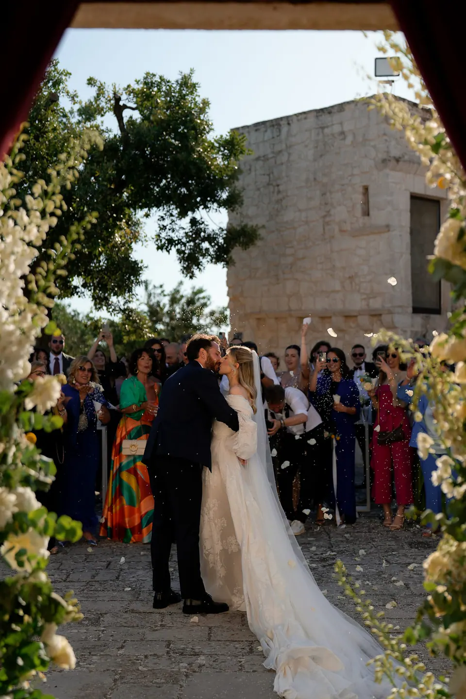 La novia recibida con un beso bajo un arco de flores blancas, rodeada de invitados.