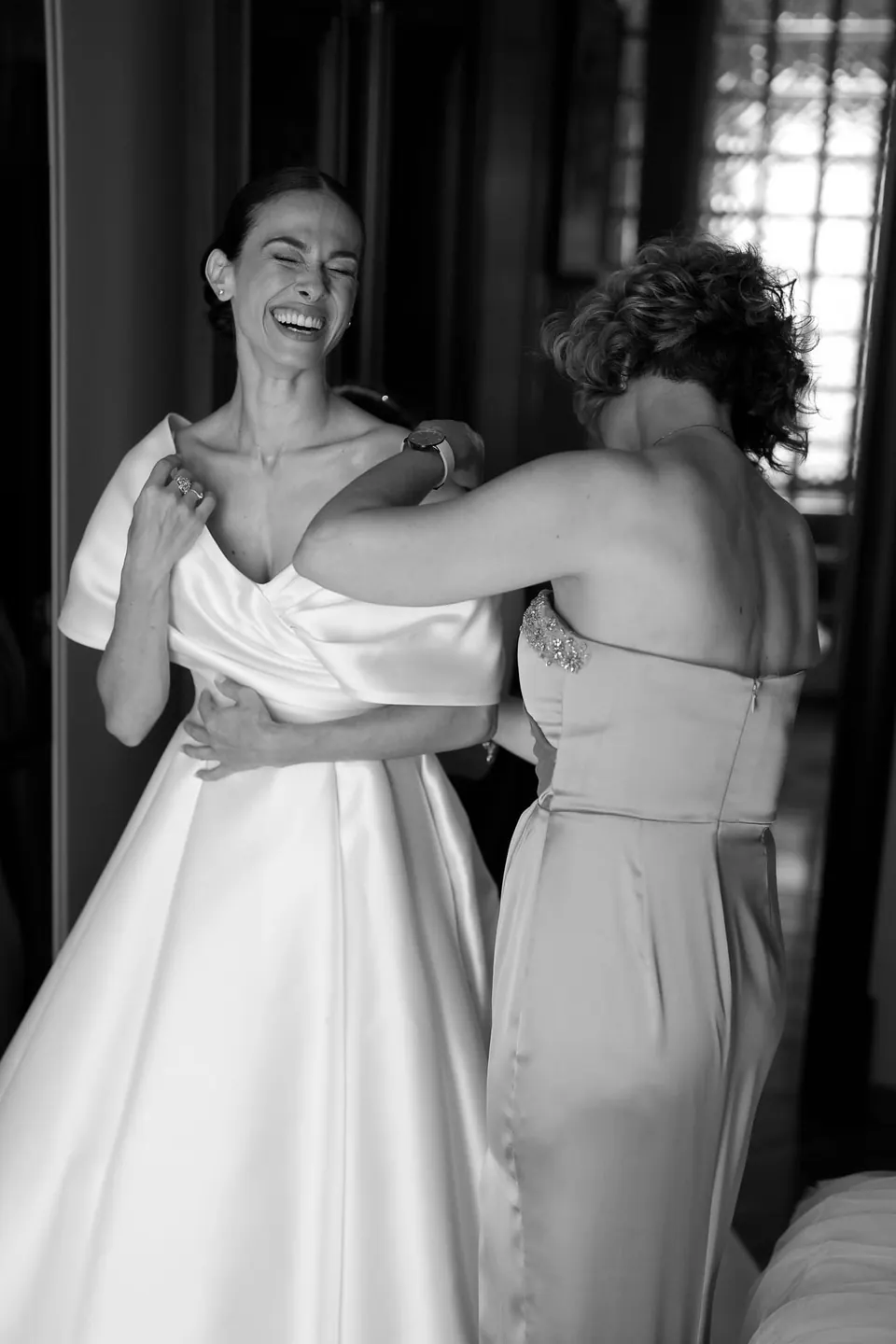 Foto in bianco e nero della sposa sorridente mentre una damigella le sistema l'abito.