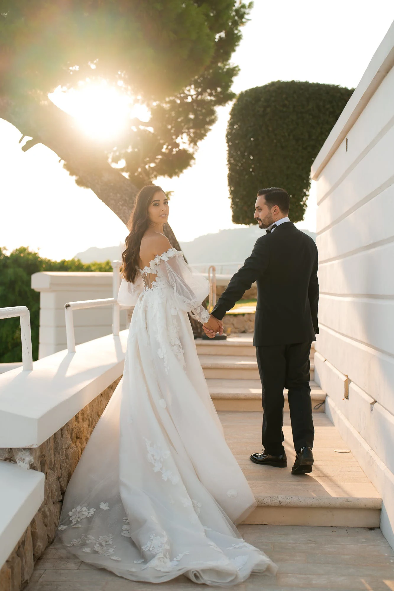 Bride and groom hand in hand on the white villa steps, sunlight through the trees.