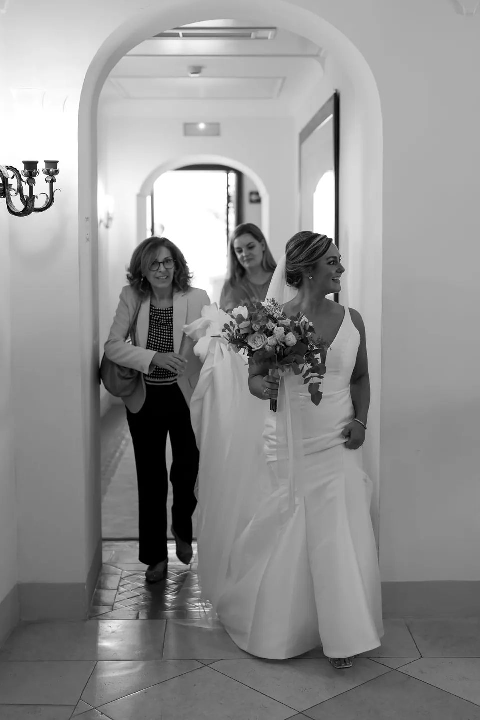 Black and white photo of the bride walking down a white corridor with the women of the family