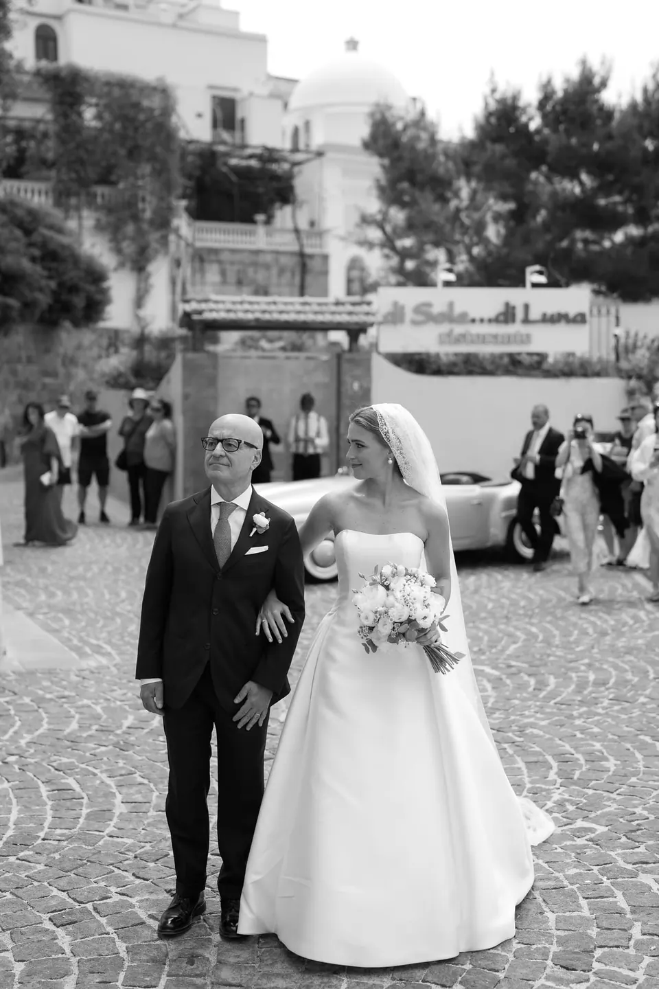 Schwarzweißfoto: Der Vater führt die Braut über die Piazza zur Kirche.