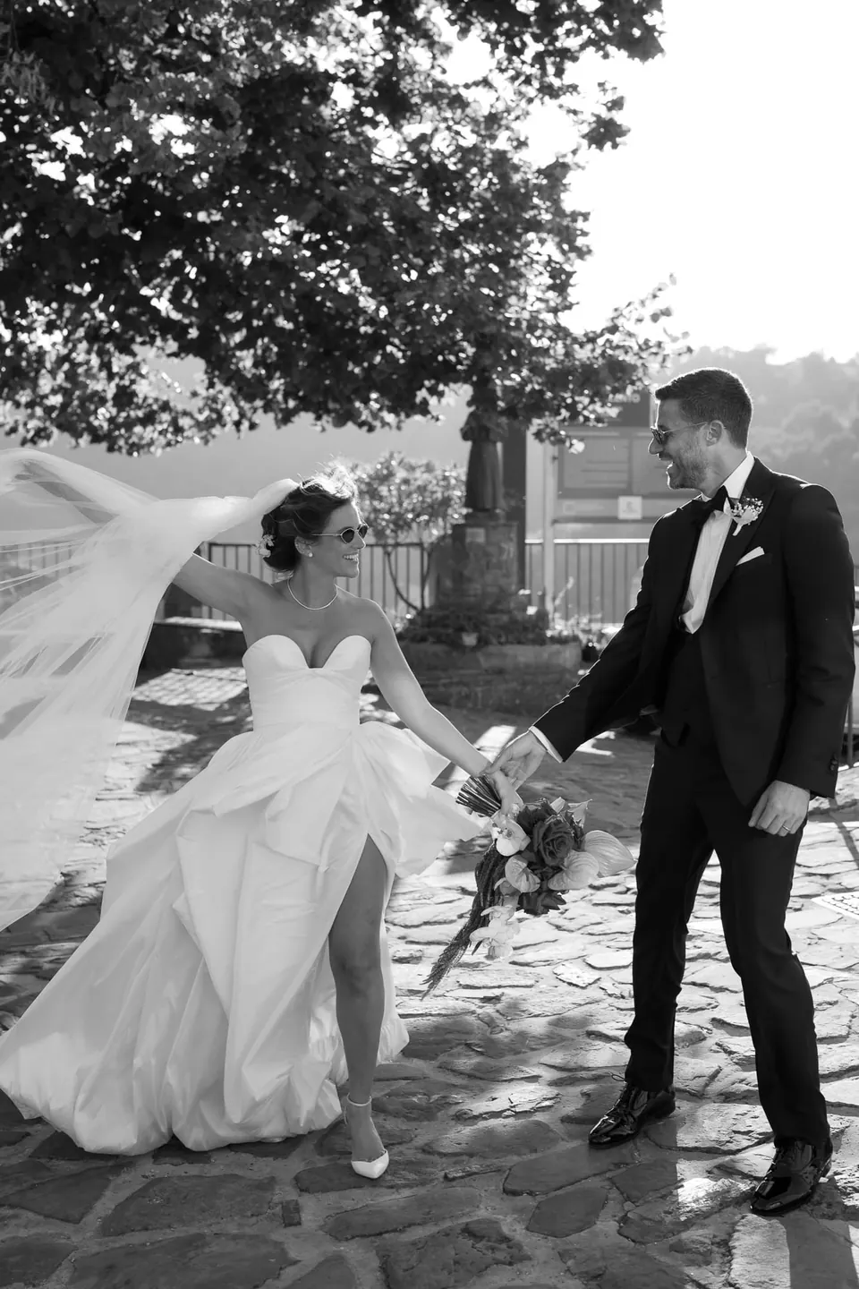 Foto in bianco e nero dello sposo che guida la sposa sul selciato, il velo che vola.