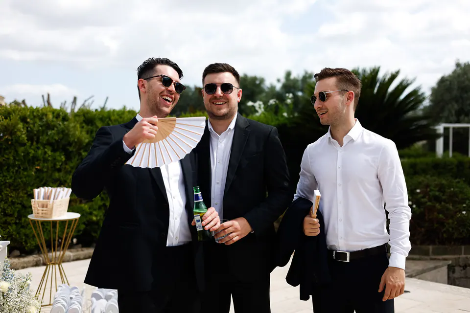 Trois amis en costume sombre et lunettes de soleil, l’un s’évente avec un éventail en papier.
