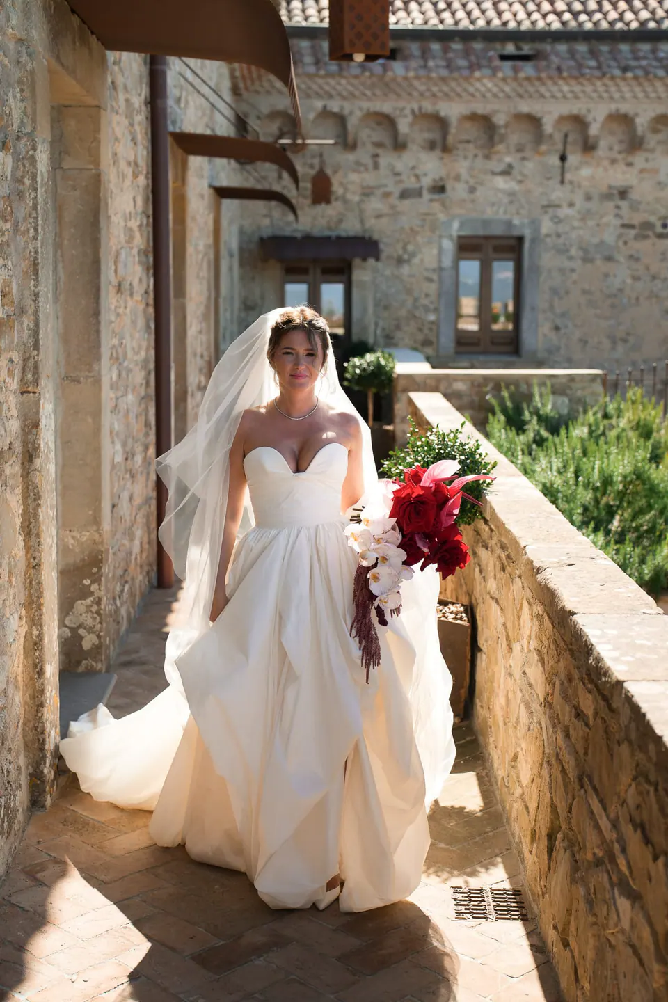 Sposa lungo un balcone in pietra assolato del castello con il bouquet rosso.