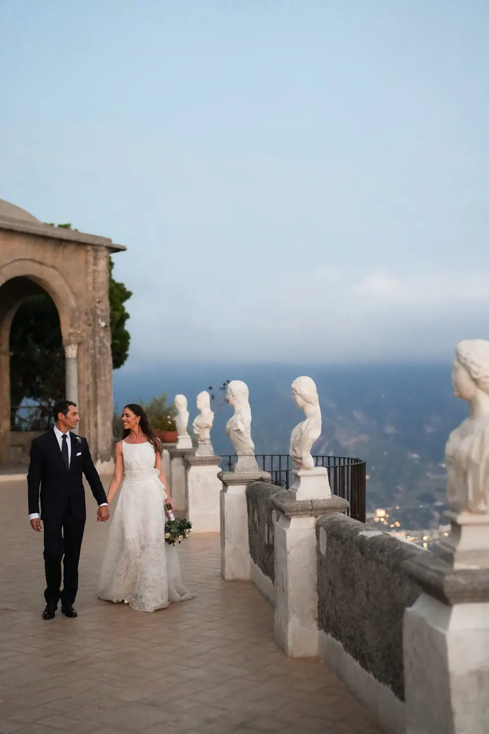 Bride and groom walking hand in hand along the Terrace of Infinity, marble busts above the clouds.
