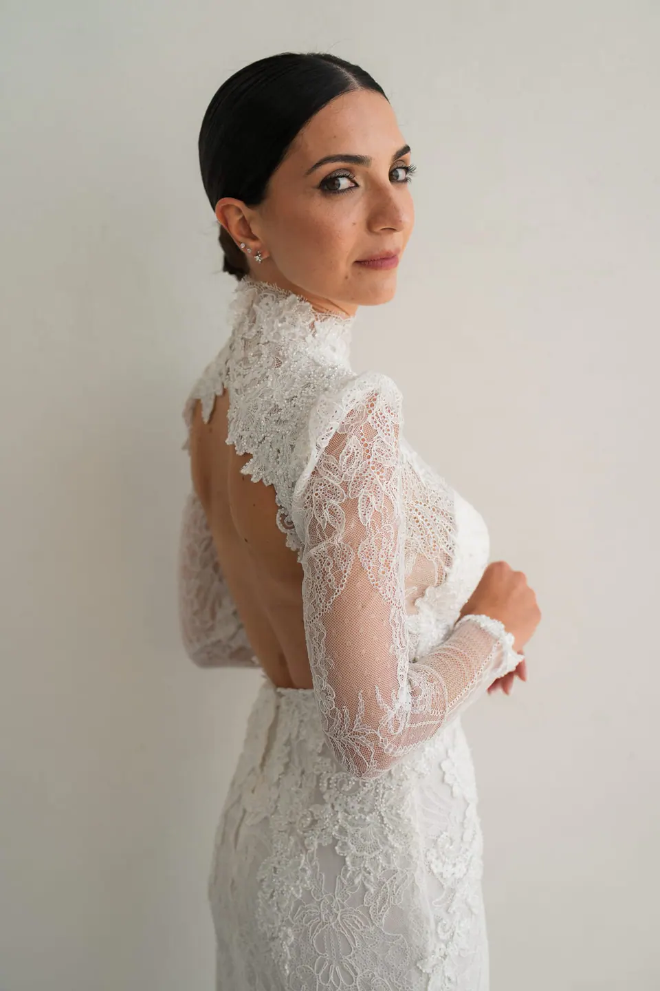 Die Braut im hochgeschlossenen Spitzenkleid mit offenem Rücken blickt über die Schulter.