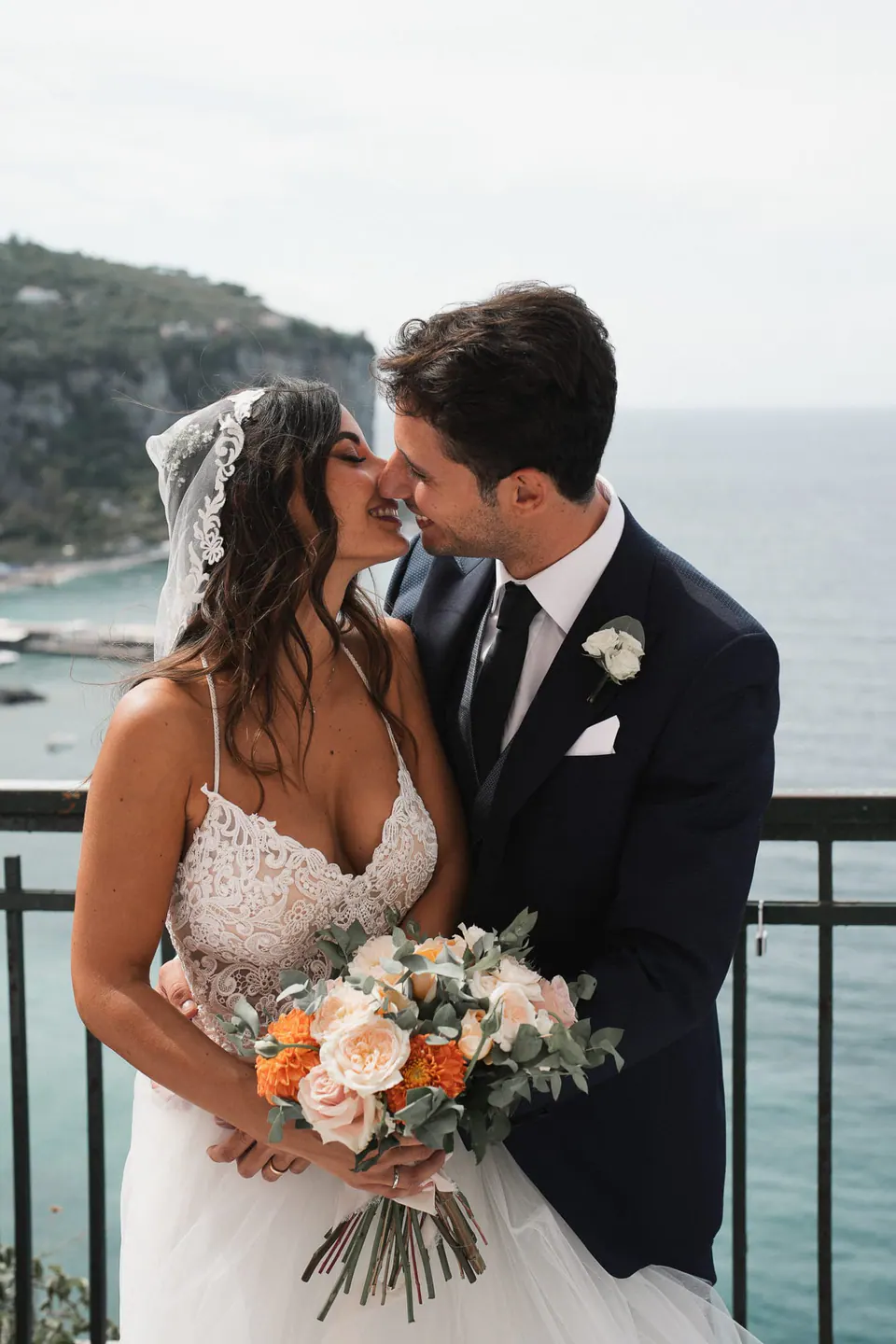 Bacio degli sposi su una terrazza sul mare a Vico Equense, il bouquet di rose pesca tra loro.