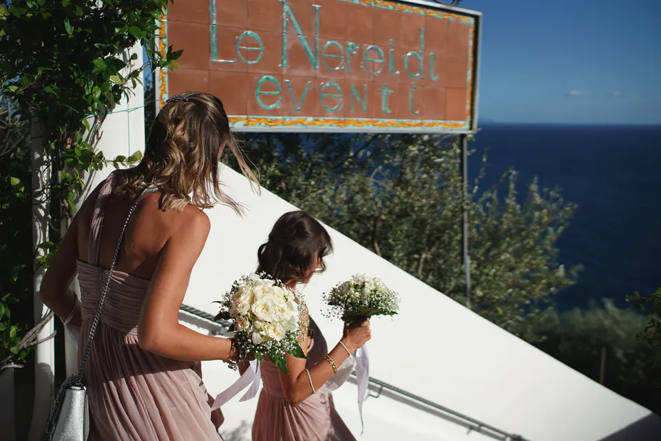 Bridesmaids with bouquets beneath the Le Nereidi sign, the sea behind them.