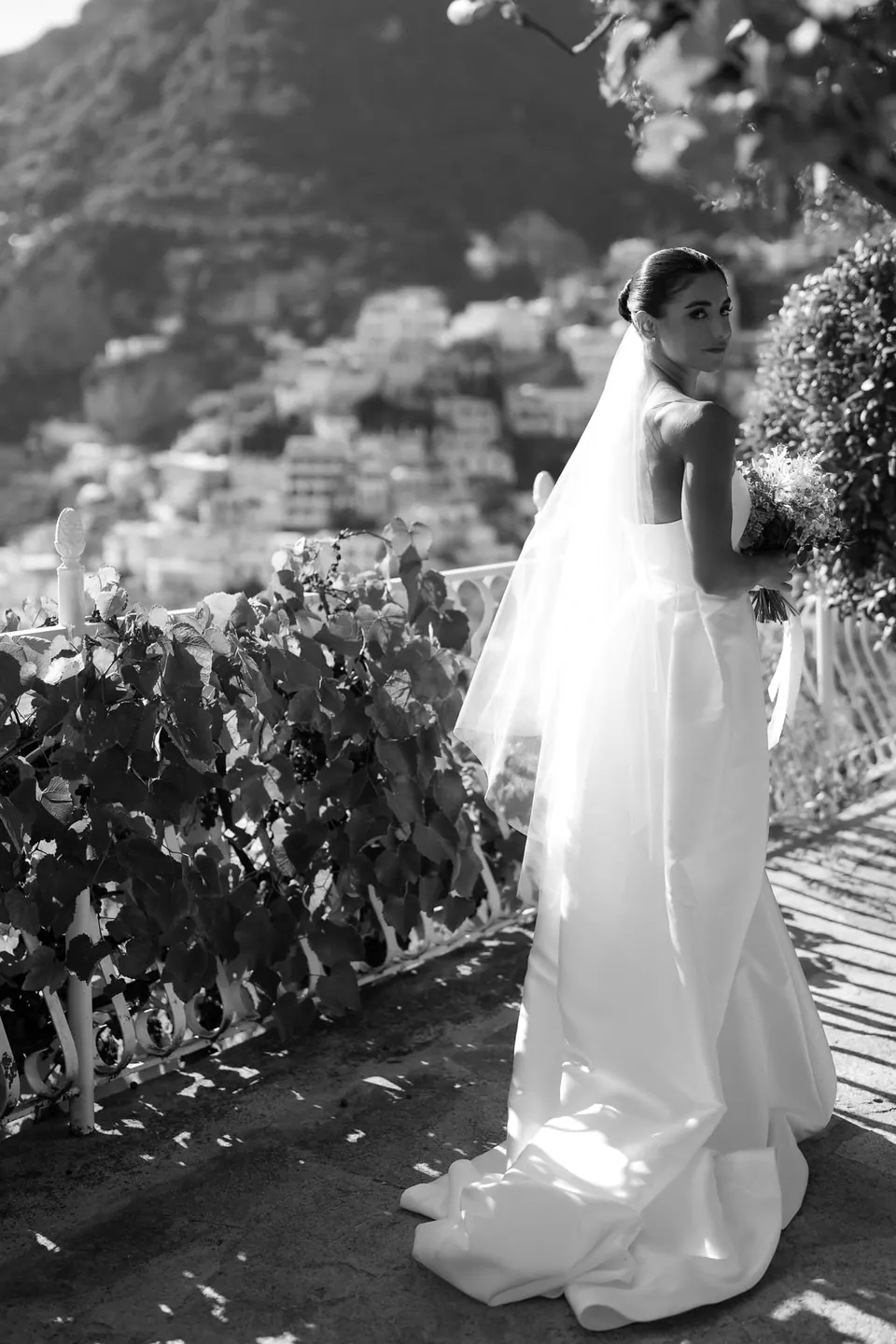 Black and white photo of the bride walking along the flowered terrace in her veil.