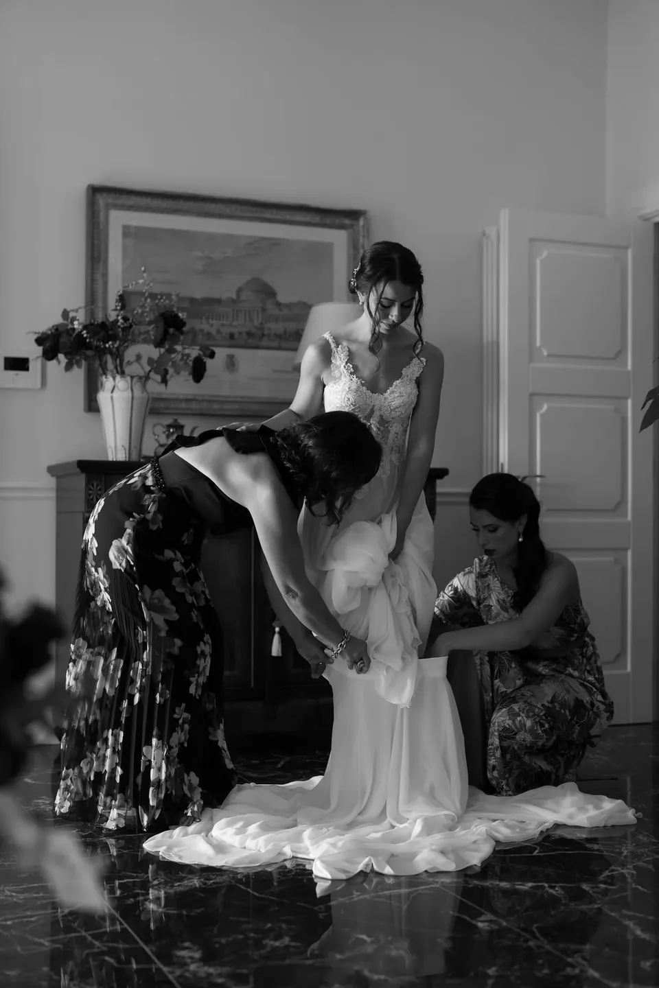 Black and white photo of women helping the bride into her gown in the bedroom.