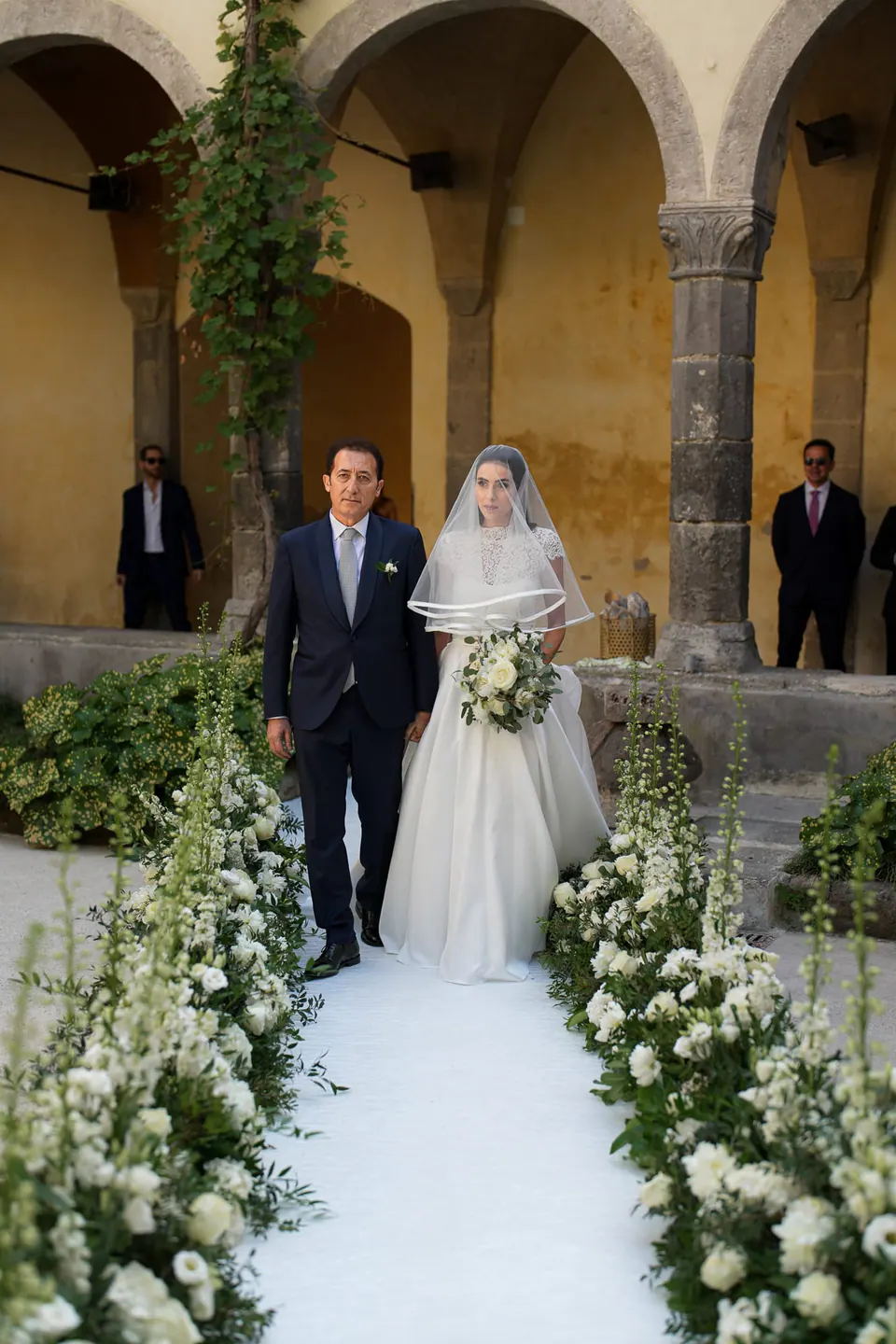 Bride walking down the flower-lined aisle in the cloister of San Francesco
