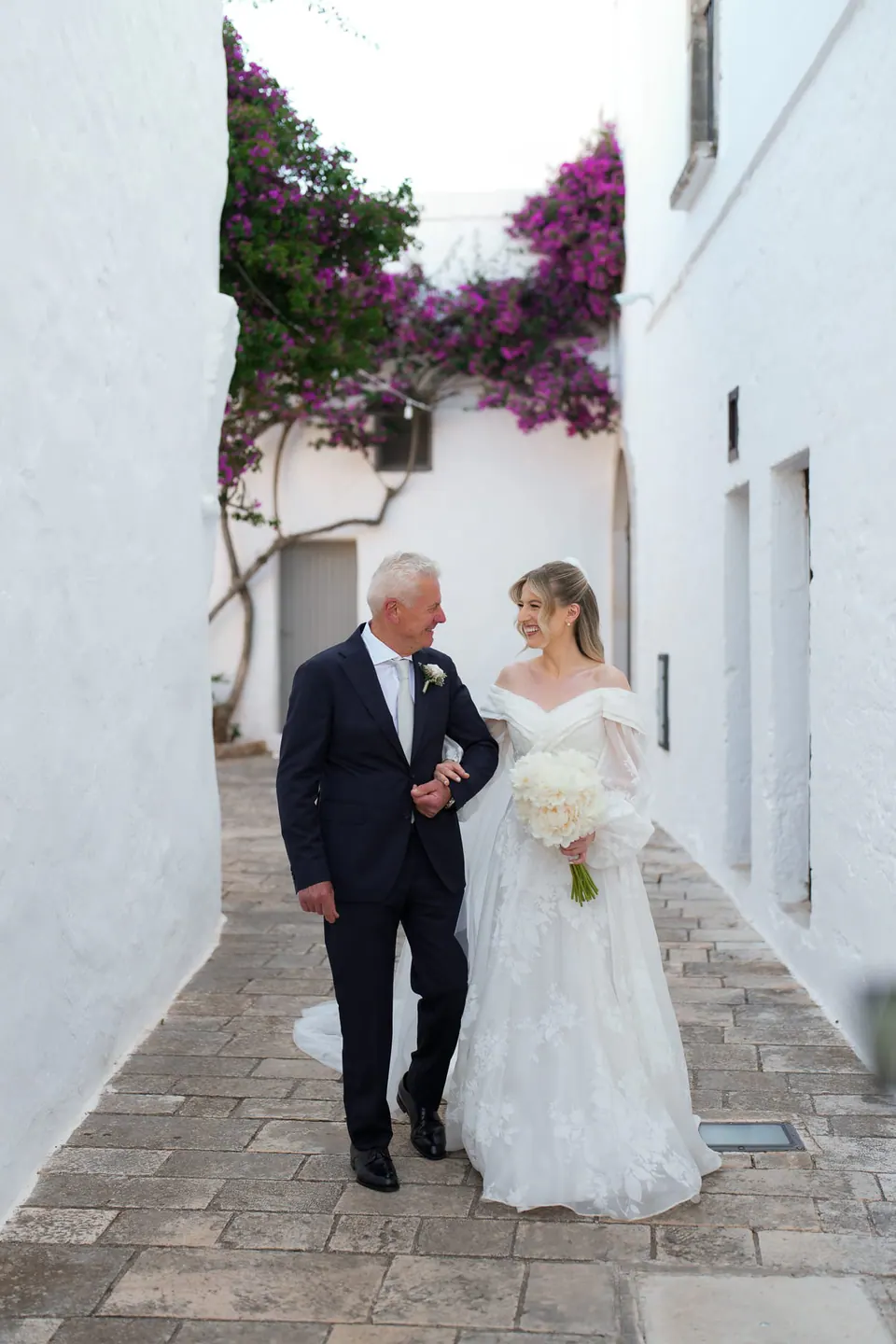La novia camina con su padre por una calle blanca y estrecha.