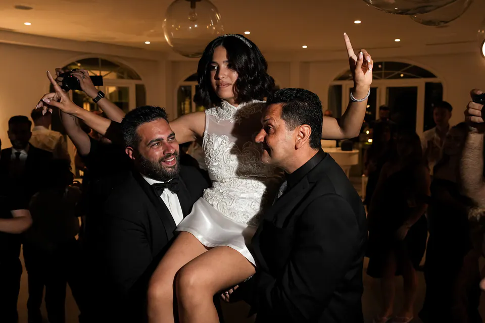 Deux invités en noir portent la mariée sur leurs épaules tandis que tout le monde danse autour.