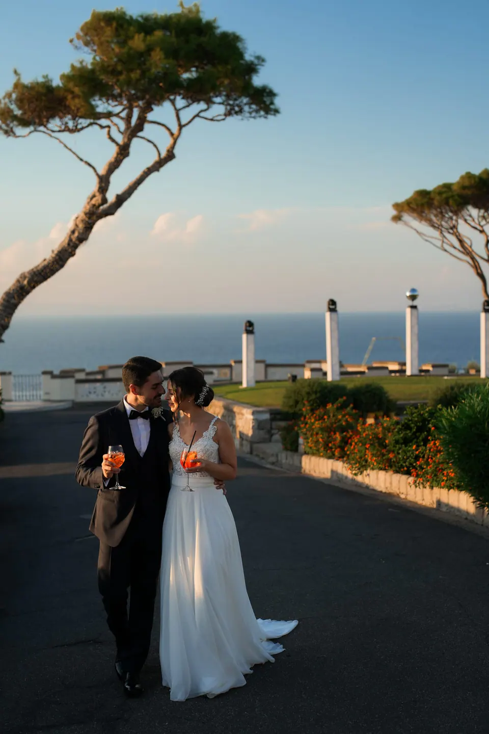 Bride and groom walking along the terrace at sunset, an umbrella pine and the sea behind them.
