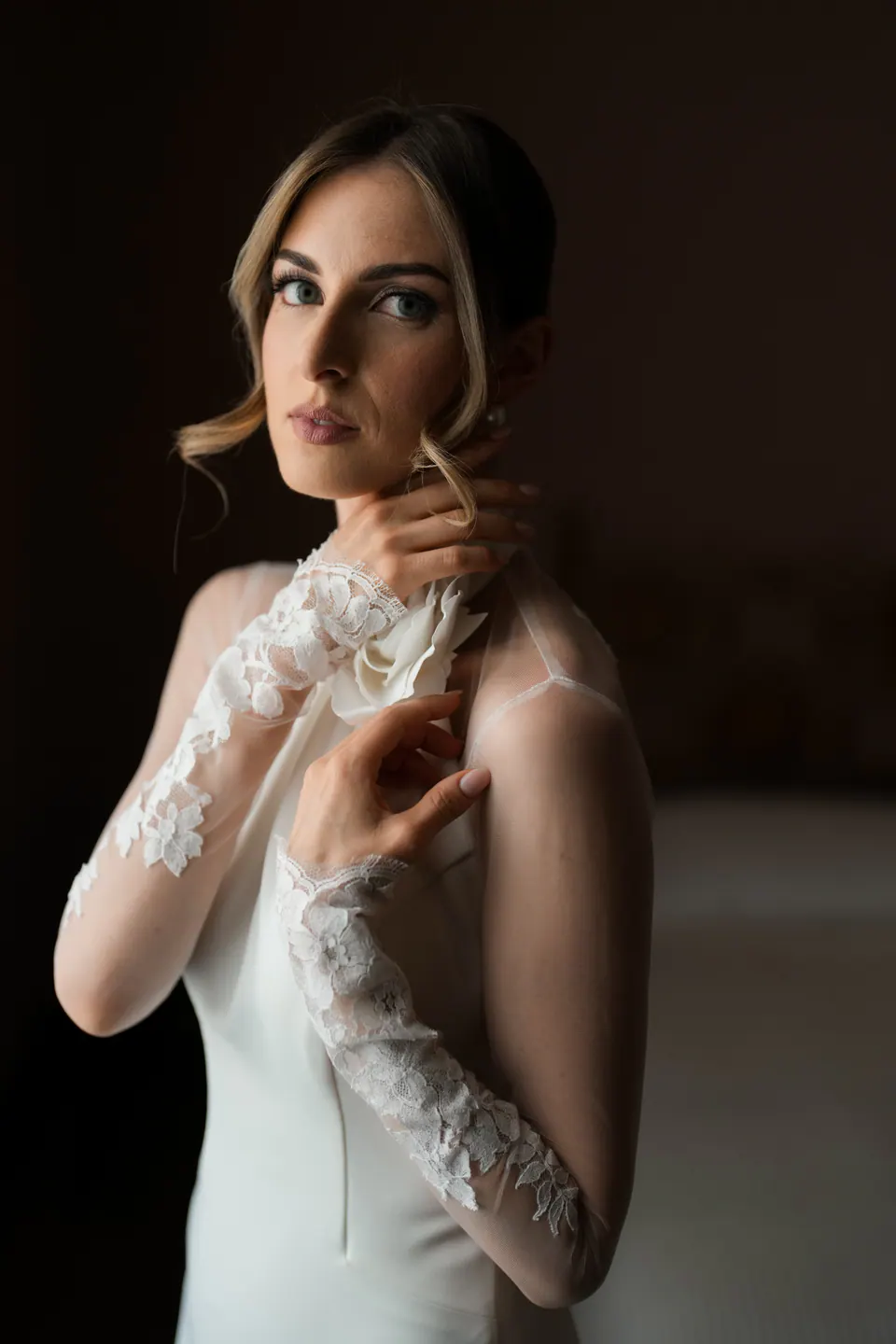 Ritratto della sposa nella luce morbida della finestra, maniche di pizzo e un fiore bianco al collo.