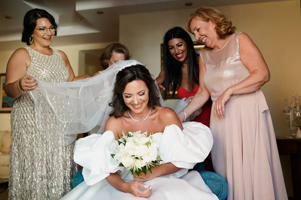 Seated bride holding her bouquet as her mother and relatives arrange her veil.