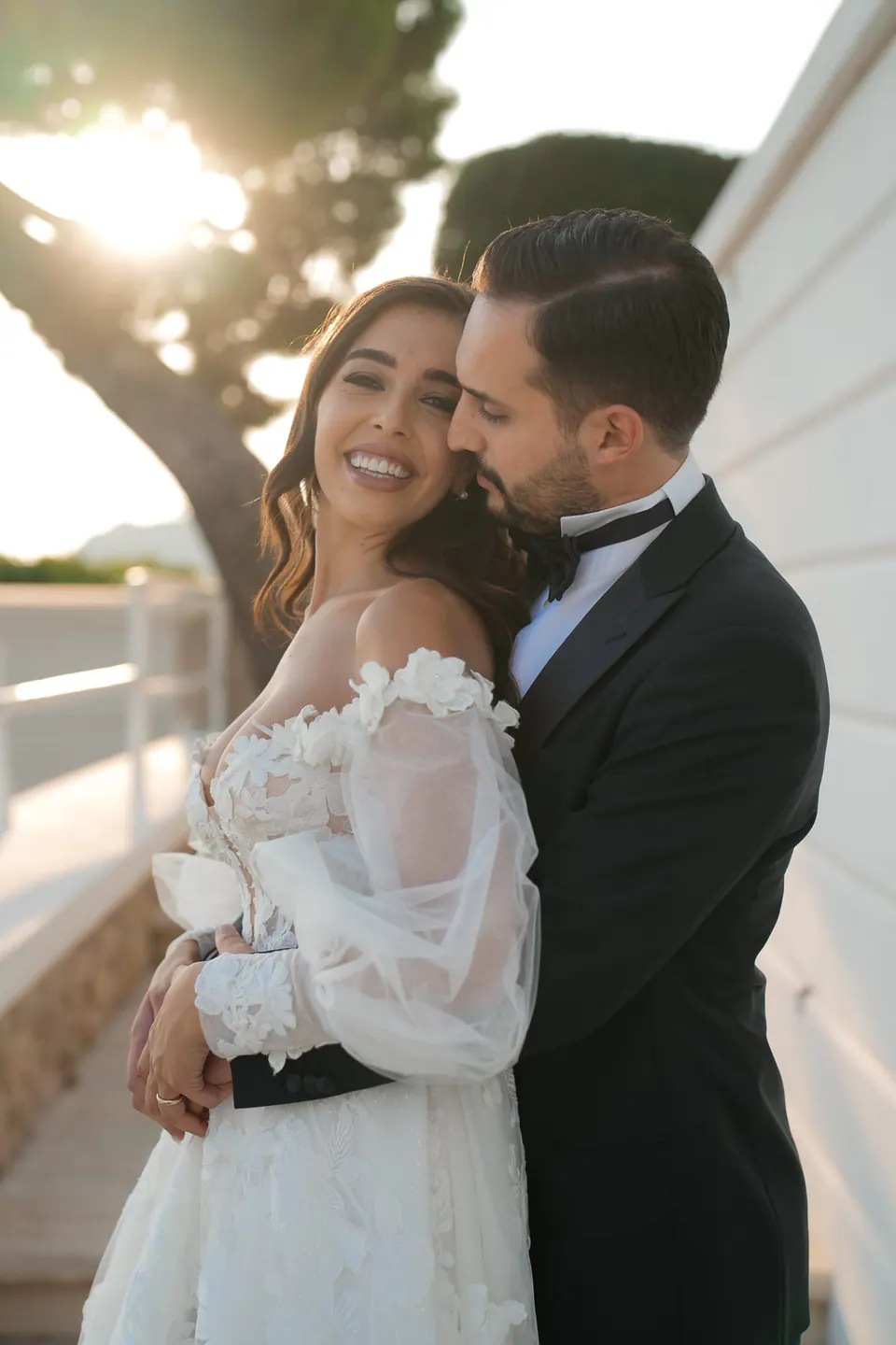 Bride and groom embracing under the coastal trees, light shimmering through the leaves.