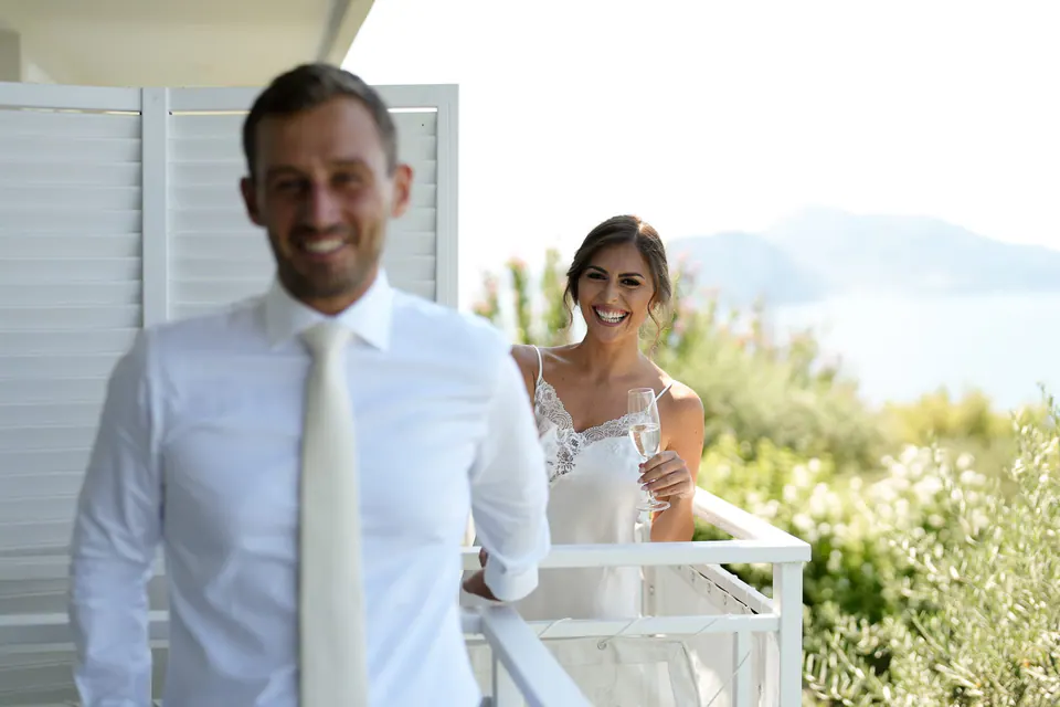Sposo sorridente in terrazza mentre la sposa aspetta sullo sfondo.