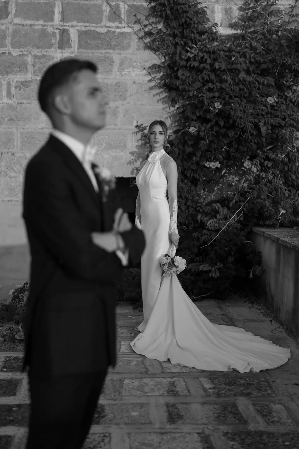 Immagine in bianco e nero con lo sposo in primo piano e la sposa dietro di lui accanto al muro d'edera.