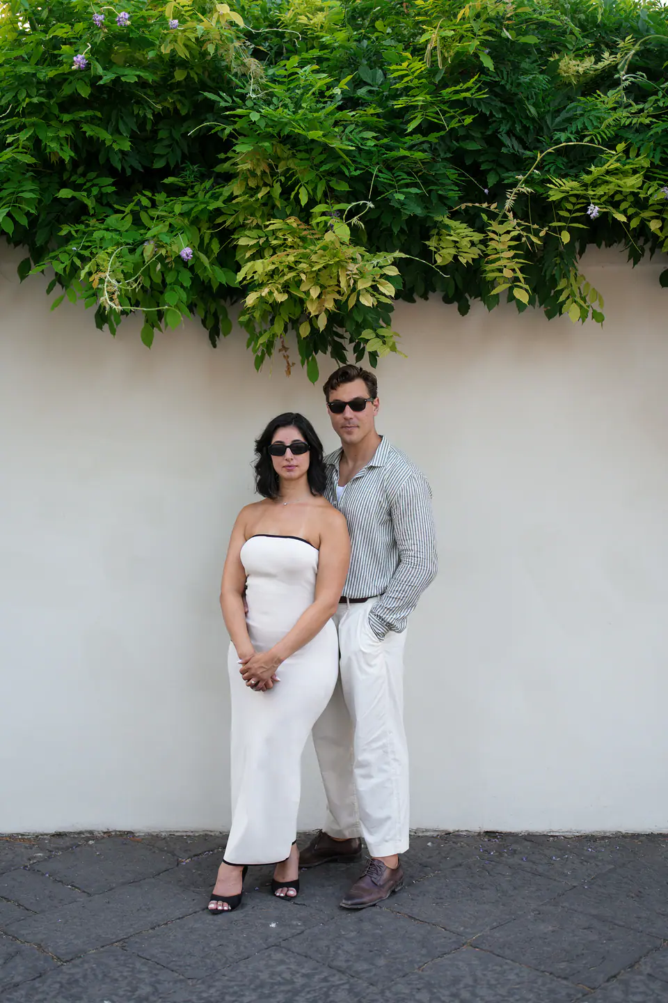 Couple standing against a white wall under green vines.