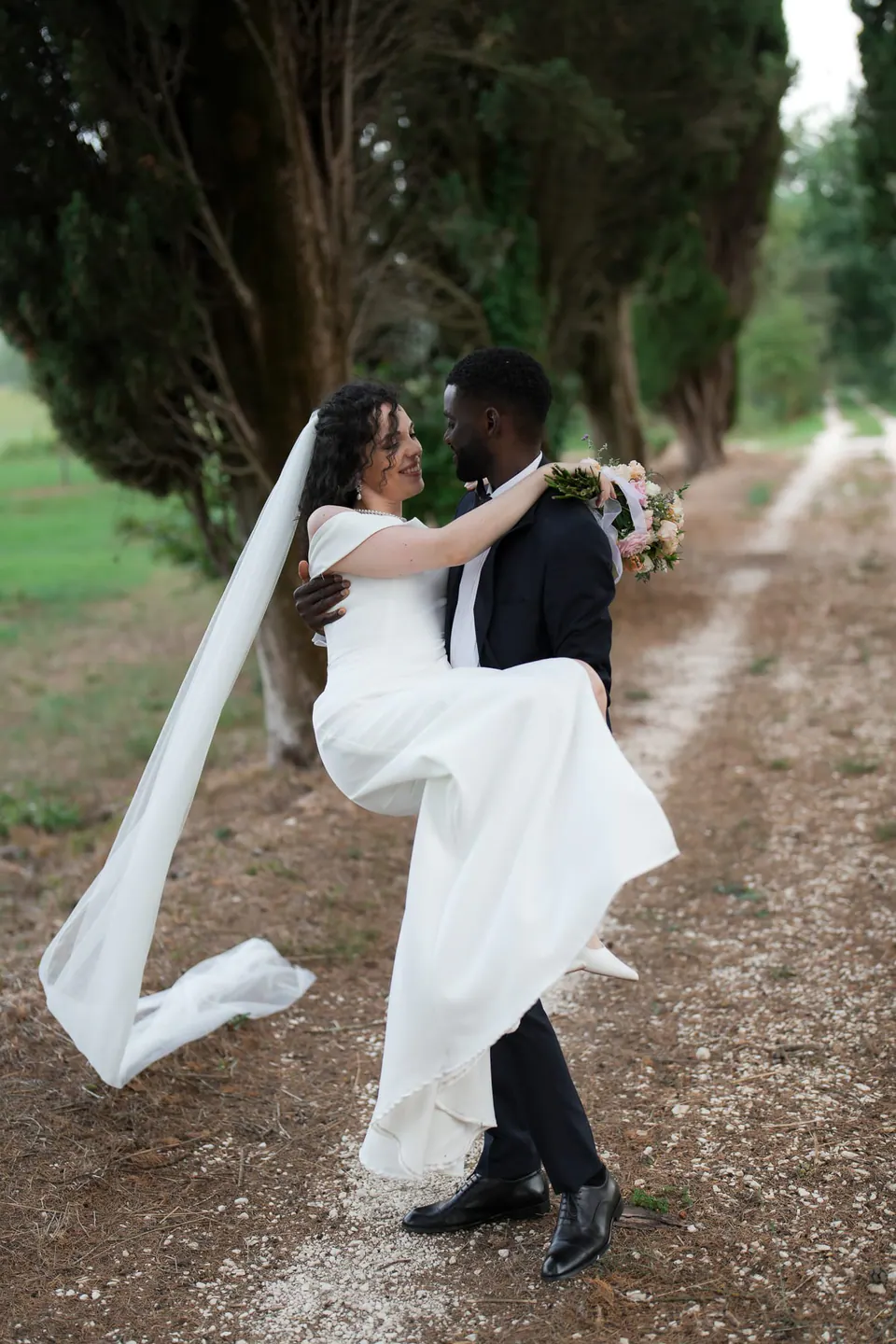 El novio levanta a la novia en un camino arbolado, con el velo volando, en Villa Ortensia D’Avalos.