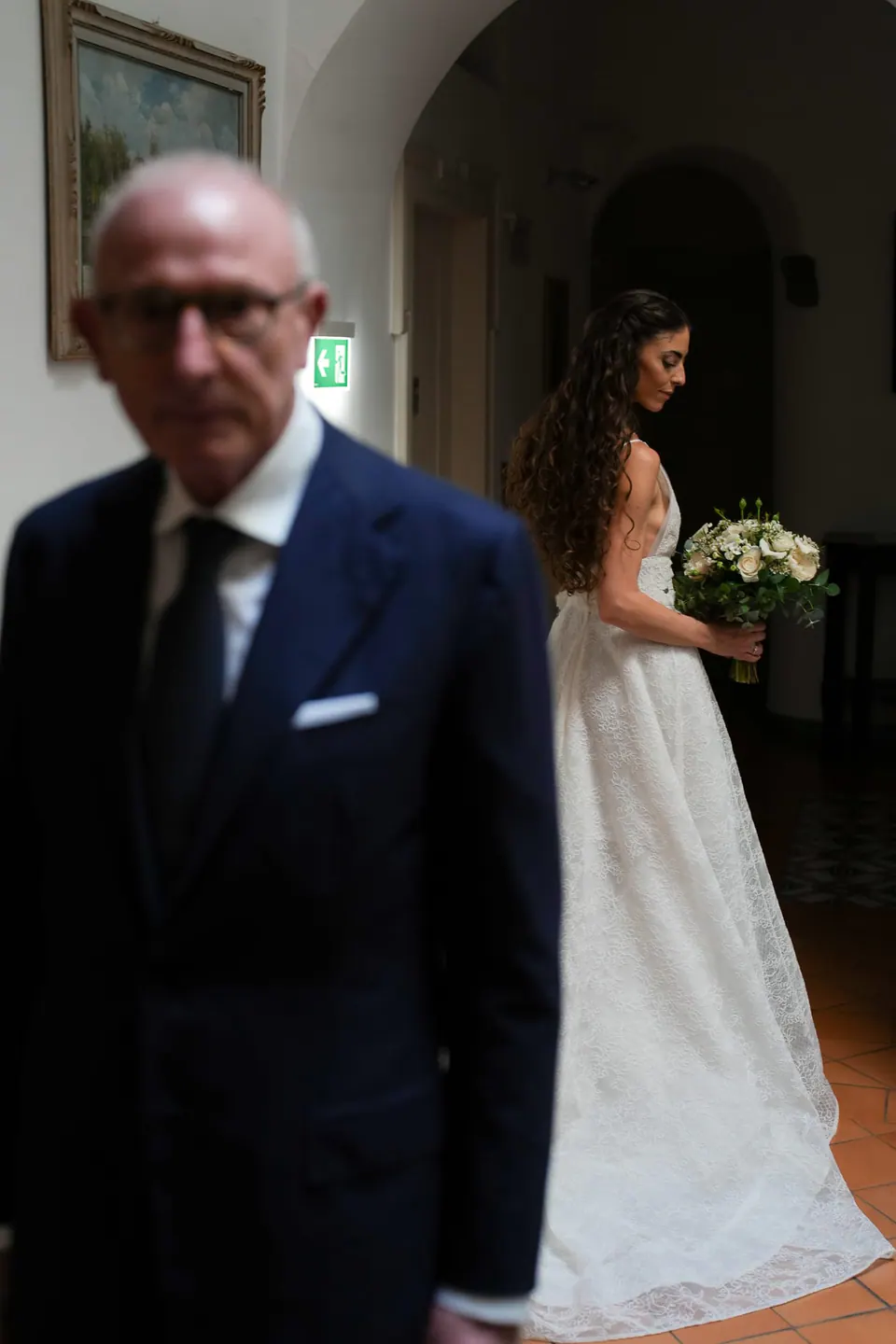 Father of the bride in the hallway as the bride walks behind him with her bouquet.