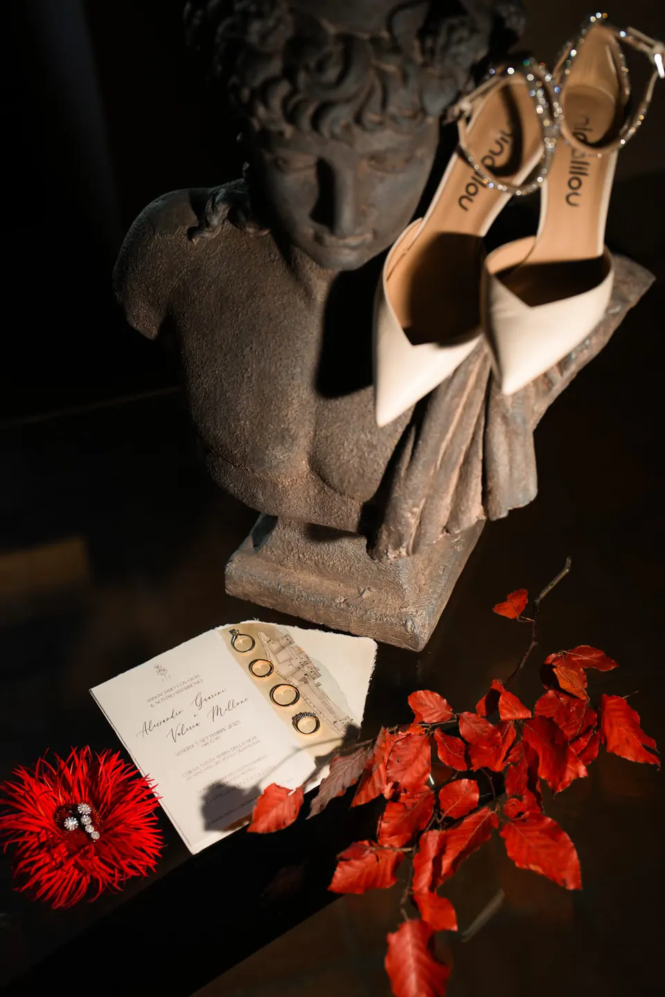 Scarpe da sposa su una statua in pietra accanto alla partecipazione e a fiori rossi, natura morta dal forte contrasto.