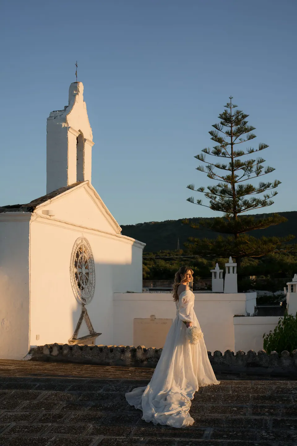 La novia delante de una pequeña capilla blanca al atardecer.