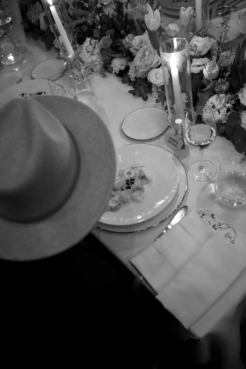 Black and white photo of a hat left on the dinner table among candles and glasses