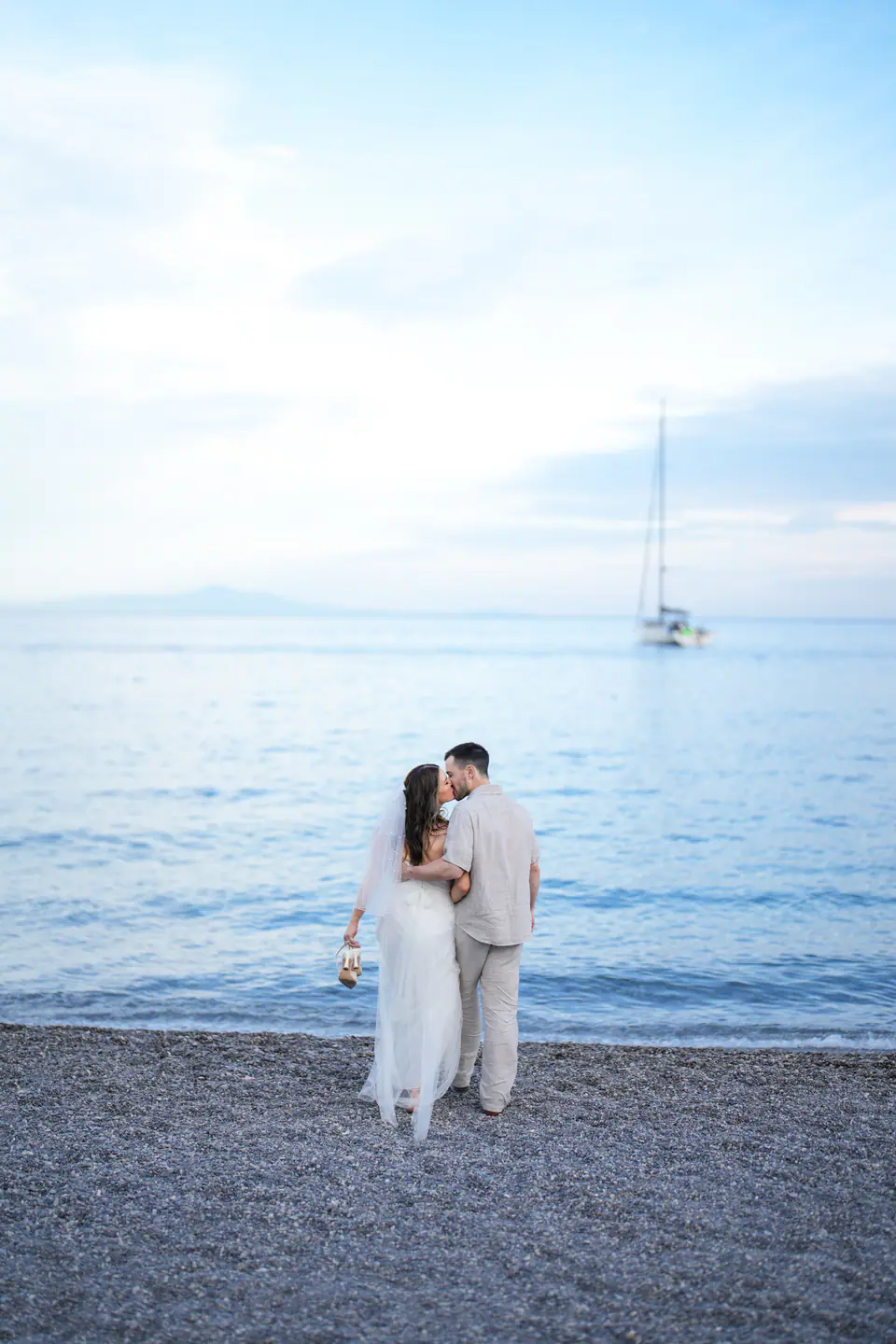 Couple standing on the empty beach at dusk, a sailing boat on the calm sea.